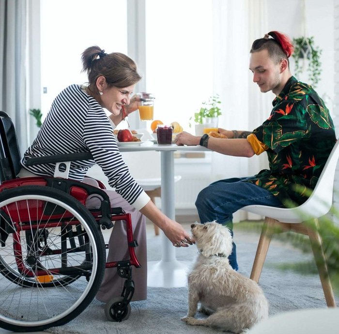 Woman using a wheelchair enjoying breakfast at home with a companion while interacting with a small dog, showing independent living and social support.