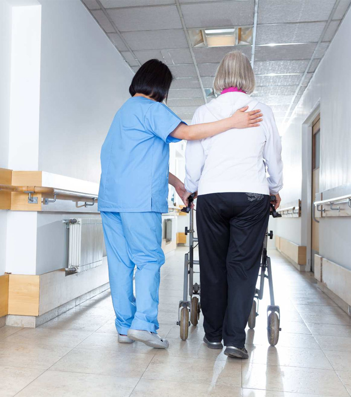 Nurse assisting an elderly woman using a walker in a hospital corridor, representing mobility support and aged care assistance services.